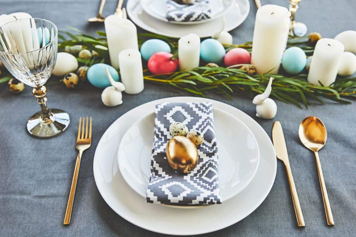 Easter table setting, white plate with gold egg on top, silverware, with greenery and colorful eggs and white candles along table