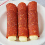 Keto Roll Ups (Pepperoni slices wrapped around string cheese) on white plate with coral and white striped napkin underneath plate, grey concrete background