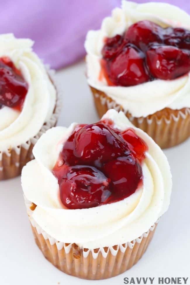 Cherry Pie Cupcake Recipe Cherry pie cupcake in front topped with pie filling and white cinnamon vanilla frosting, 2 cherry cupcakes behind and purple cloth in the background.