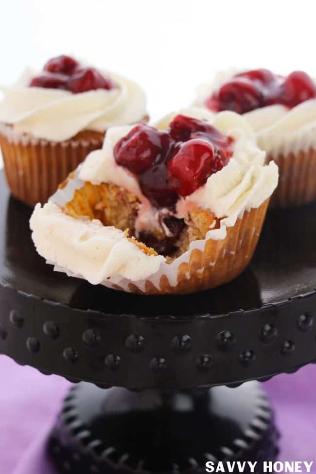 Easy Cherry Pie Cupcakes With Cinnamon Vanilla Frosting Cherry cupcake with a bite taken out with cherry pie filling coming out and topped with frosting. Two other cherry pie dessert cupcakes in the background, all on a black cake stand.