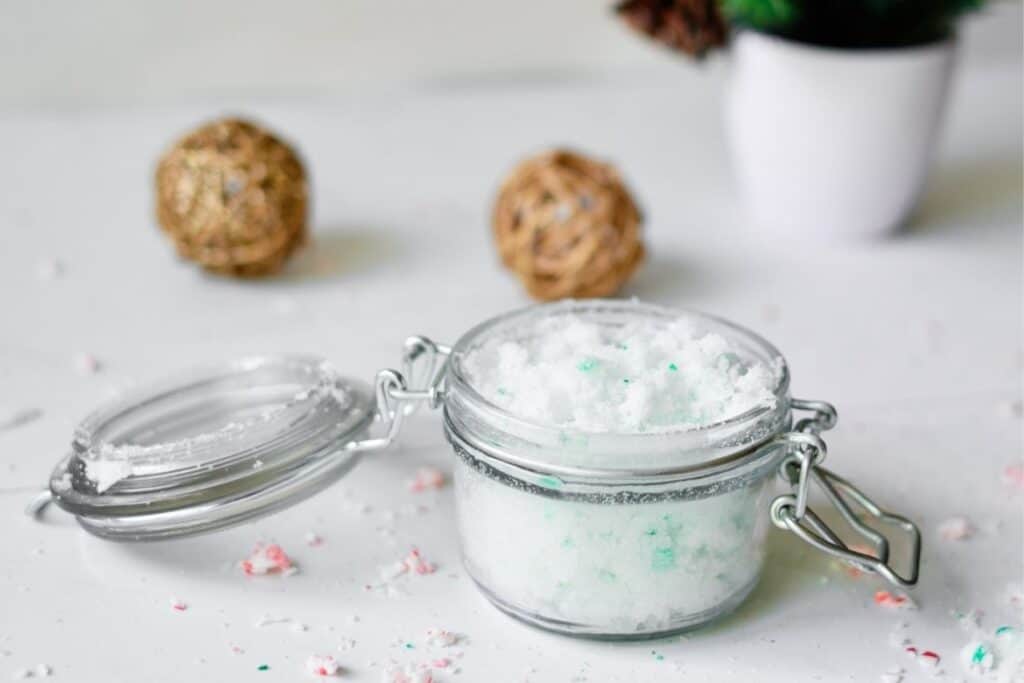 Christmas peppermint sugar scrub in a clear container with peppermint candies sprinkled in the background. Also the bottom of a Christmas tree and wood balls behind the DIY sugar scrub.