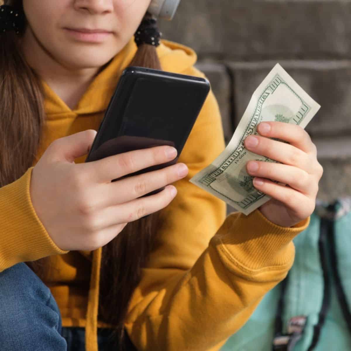 Girl with brown hair in a gold sweatshirt holding up her phone in one hand and hundred dollar bill in the other hand, turquoise backpack and brick wall in the background.
