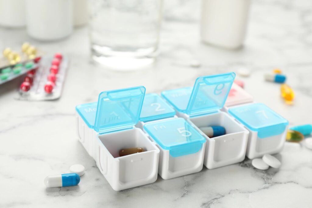 A weekly pill organizer with blue lids and white compartments, some containing colorful pills and capsules, sits on a marble countertop with medical supplies in the background.
