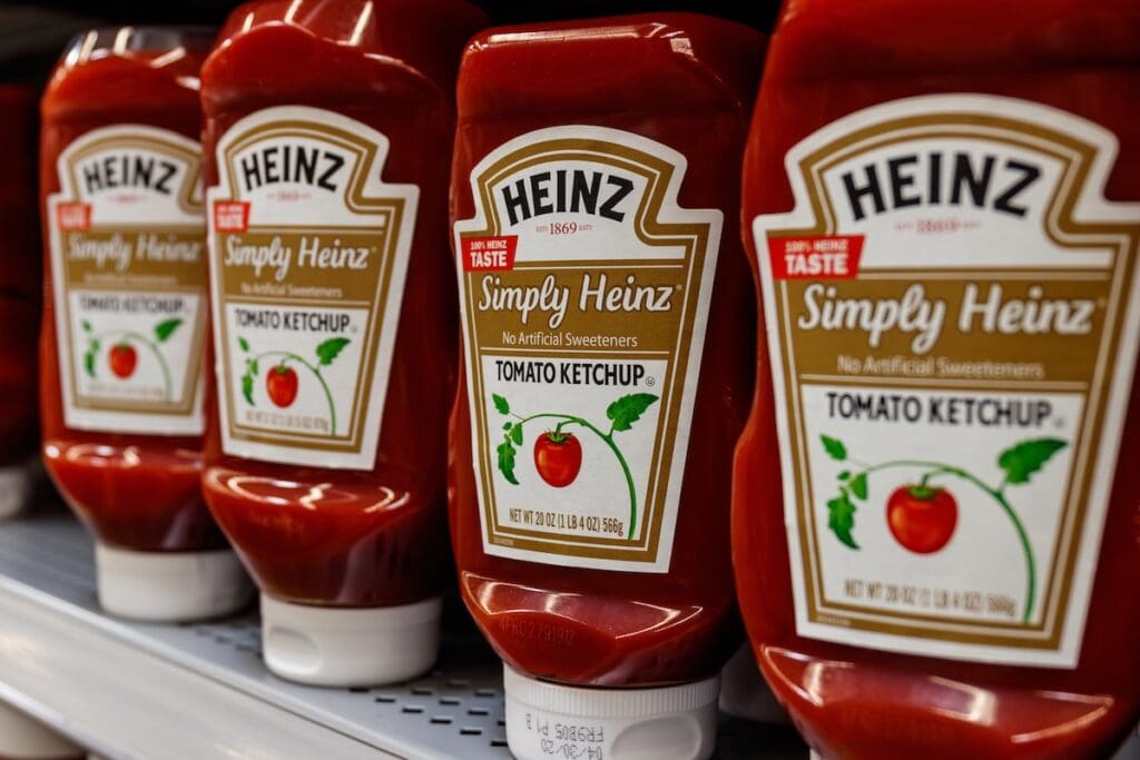 Heinz Simply Heinz tomato ketchup bottles arranged on a store shelf, featuring the classic red squeeze bottles with white caps and labels showing a tomato graphic and "No Artificial Sweeteners" messaging.