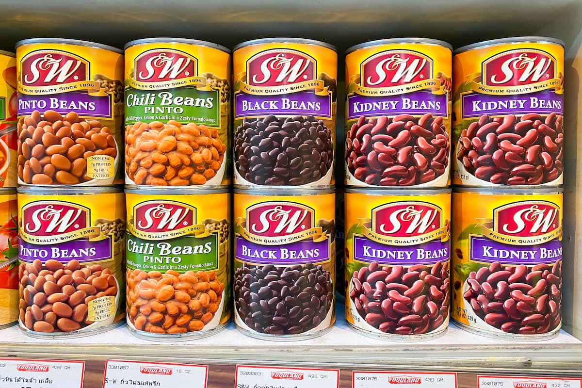 Rows of S&W brand canned beans displayed on a grocery store shelf, including varieties of Pinto Beans, Chili Beans, Black Beans, and Kidney Beans in yellow and orange labeled cans with price tags below.