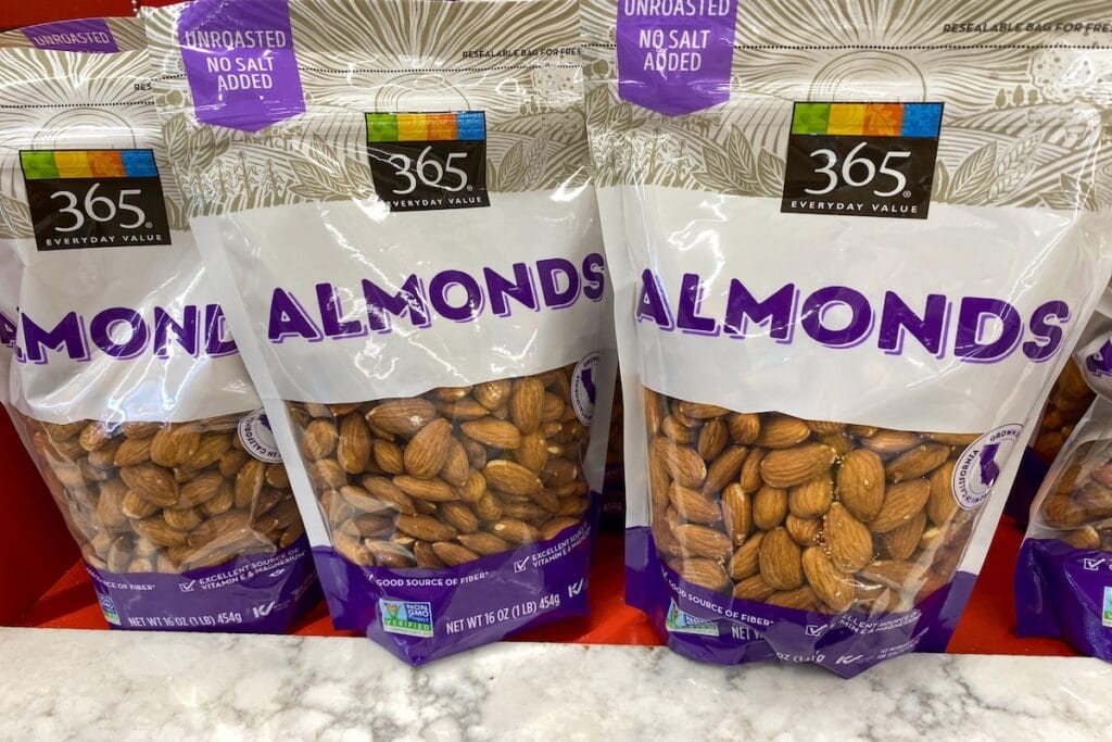 Three bags of 365 Everyday Value brand almonds on a store shelf, featuring clear packaging with purple and white labeling that indicates "Unroasted No Salt Added" with visible whole almonds inside each resealable bag.