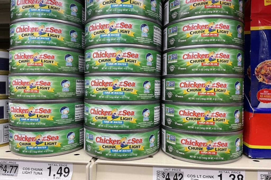 Stacks of Chicken of the Sea brand chunk light tuna cans arranged on store shelves, showing both "Tuna in Water" and "Tuna in Oil" varieties in green and yellow packaging with price labels displaying costs like $1.49.
