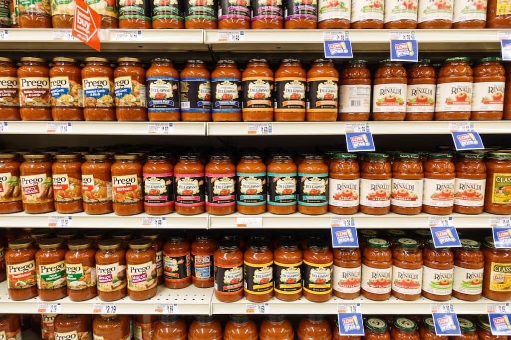Grocery store shelves displaying multiple brands of pasta sauce including Prego, DelGrosso, and Rinaldi varieties in glass jars, organized in rows with blue promotional price tags and various sauce types like marinara and meat sauce.