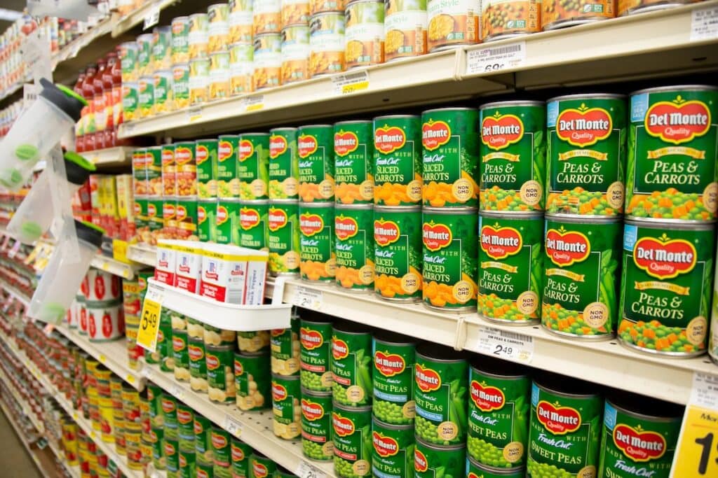 Multiple shelves of Del Monte canned vegetables in a grocery store aisle, prominently featuring green cans of diced carrots, sliced carrots, peas and carrots, and sweet peas with price tags and promotional signs visible.