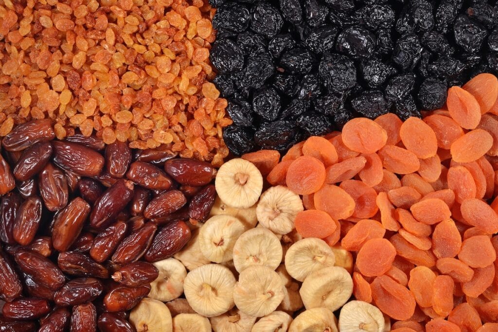 An assortment of dried fruits arranged in groups including golden raisins, dark raisins, dates, dried apricots, and dried banana chips displayed on a white surface.