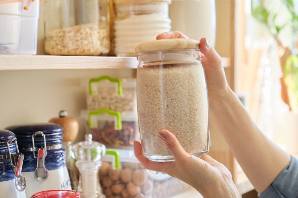  Hands holding a glass storage jar filled with what appears to be rice or another grain, with additional storage containers visible on wooden shelves in an organized pantry.
