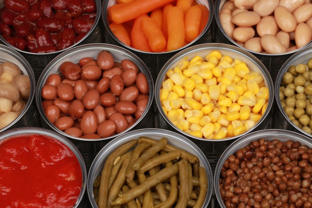 An overhead view of multiple metal bowls containing various canned and preserved foods including kidney beans, corn, white beans, pickles, tomato sauce, and other pantry staples arranged in a grid pattern.