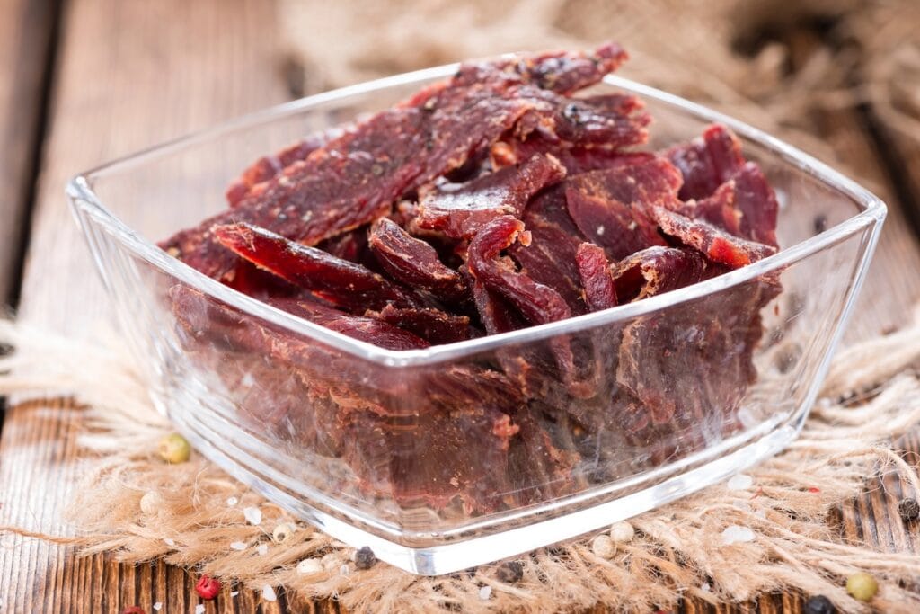 A clear glass bowl containing dark red beef jerky strips sits on a rustic wooden surface with burlap fabric visible in the background.