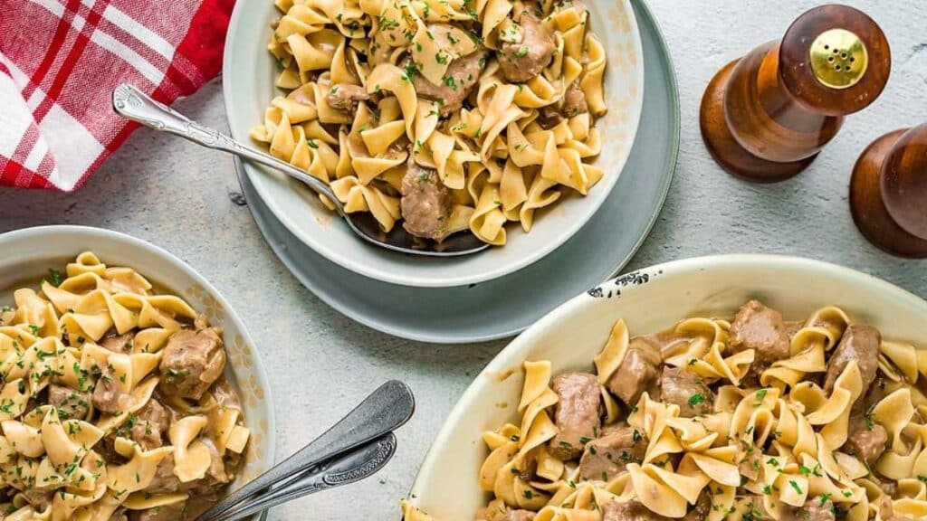 Three bowls of slow cooker beef and noodles with mushrooms on a table with spoons in them.