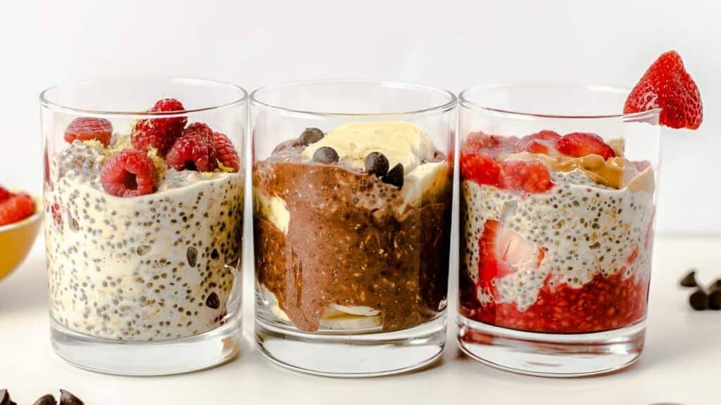 Overnight oatmeal in glasses topped with fruit, chocolate, and chia seeds on a light background.