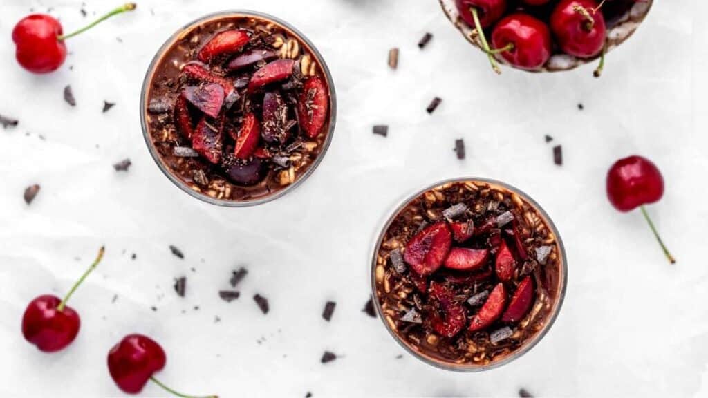 Chocolate Cherry Overnight Oats in jars next to cherries and chocolate chunks on a marble background. 
