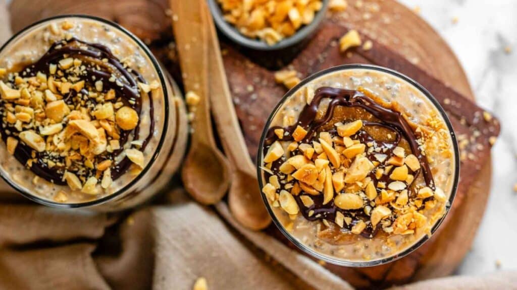 Snickers overnight oats in jar topped with date caramel, chocolate and chopped peanuts on a wood cutting board. 