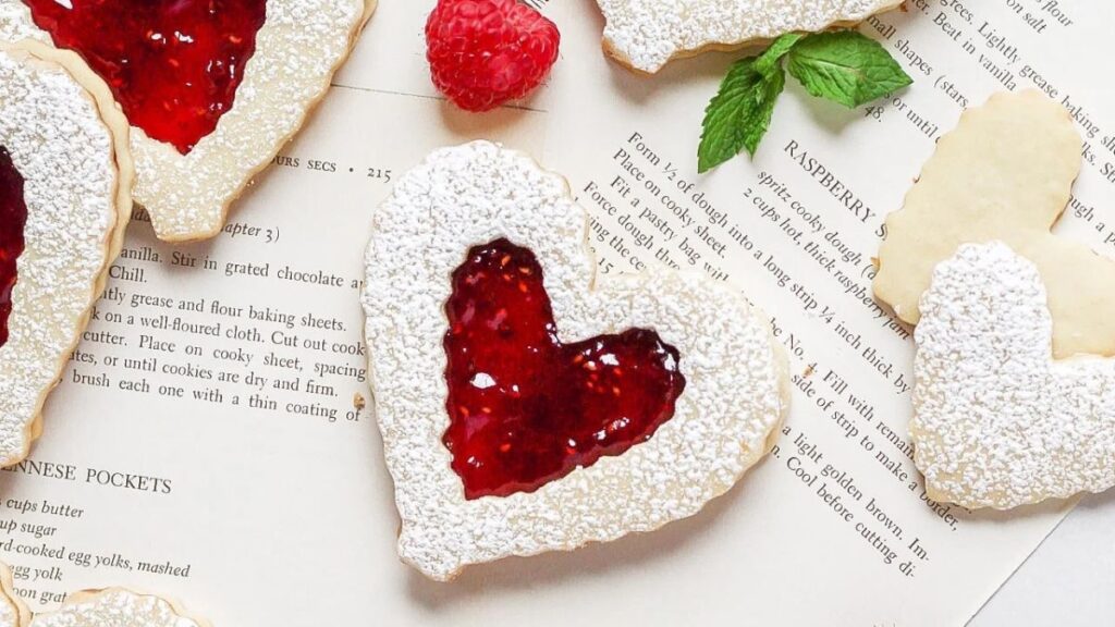 Heart-shaped raspberry Linzer Cookies with raspberries nearby.
