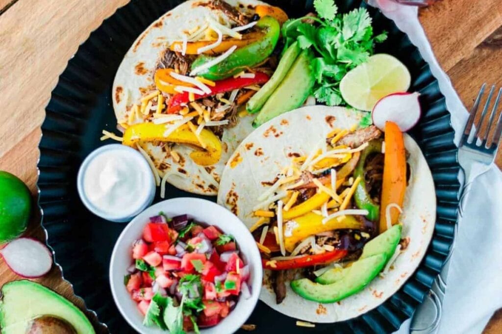 Pork carnitas fajitas on a plate with a bowl of sour cream and toppings.