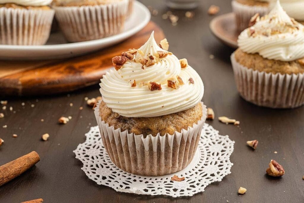 A single cupcake with white frosting and pecan pieces sits on a doily on a dark brown surface. More cupcakes and pecan pieces are visible in the background. A cinnamon stick is in the foreground.