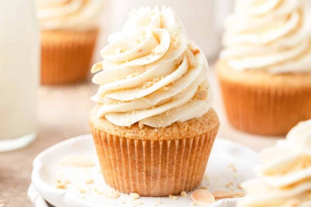 A single cupcake with vanilla frosting and almond pieces is in focus on a white plate. Other similar cupcakes are blurred in the background. A glass of milk is partially visible in the far left background.