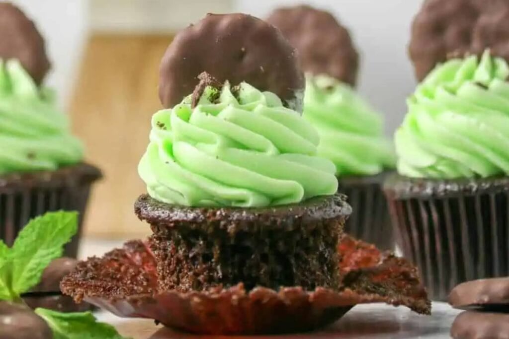 A chocolate cupcake with mint green frosting and a chocolate mint on top is in focus. Other similar cupcakes are blurred in the background. Mint leaves and chocolate mints are visible on the table.