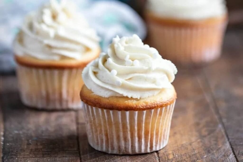 Three vanilla cupcakes with vanilla frosting and white sprinkles are on a wooden surface. Another cupcake is blurred in the background.