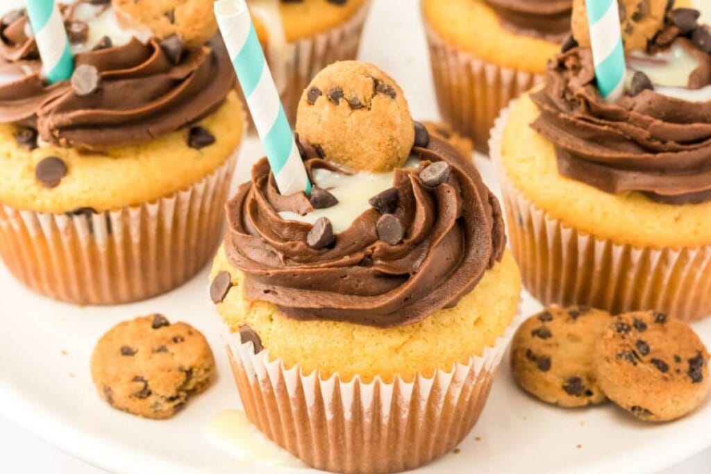 Close-up of several cupcakes with chocolate frosting, chocolate chips, and a mini chocolate chip cookie on top. A small white plate is underneath. Some extra cookies are scattered around the cupcakes. A few cupcakes have a striped straw stuck in them.