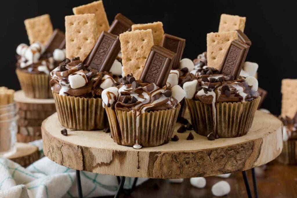 Several chocolate cupcakes are arranged on a rustic wooden cake stand. Each cupcake is topped with chocolate frosting, marshmallows, chocolate chips, and a graham cracker. More cupcakes and graham crackers are visible in the background.