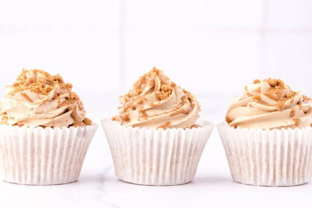 Three cupcakes with tan frosting and Biscoff cookie crumbles are arranged in a row on a white surface.