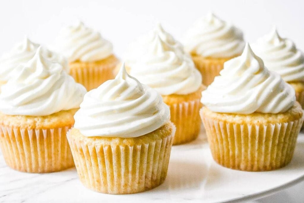 Several cupcakes with tall swirls of white frosting are arranged on a white surface.