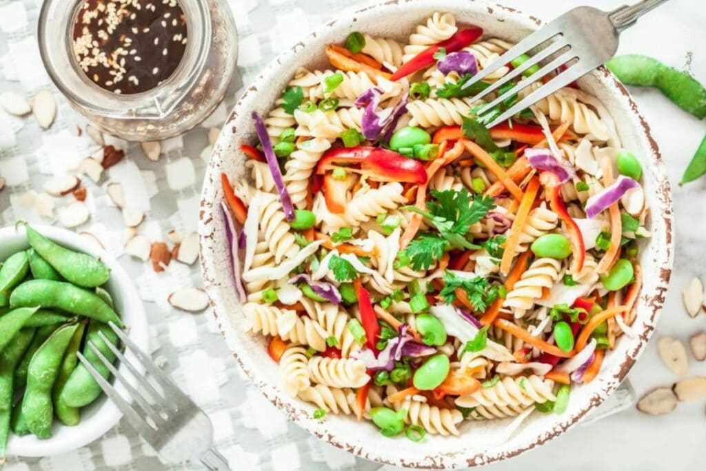 A bowl of pasta salad with rotini pasta, edamame, shredded carrots, red cabbage, and bell peppers. A small bowl of edamame and a jar of dressing are in the background.