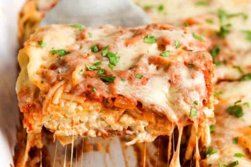 A spatula lifts a serving of baked spaghetti casserole. The casserole is topped with melted cheese and chopped parsley. Strings of cheese are stretching from the serving.
