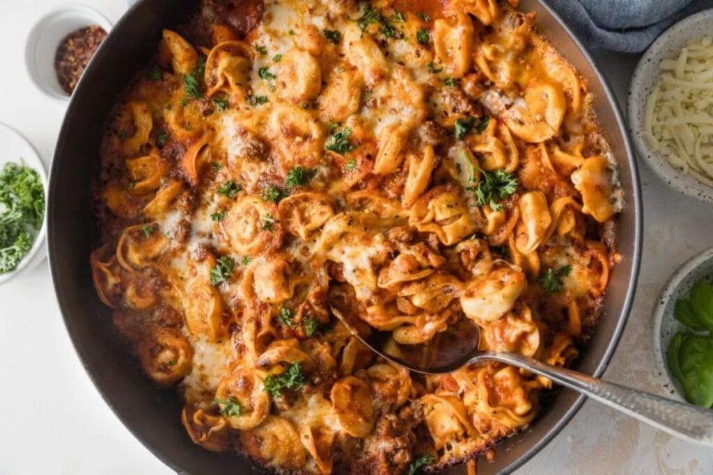 A skillet of baked tortellini. The tortellini is covered in a meat sauce and melted cheese. Fresh parsley is sprinkled on top. A spoon is taking a serving from the skillet.
