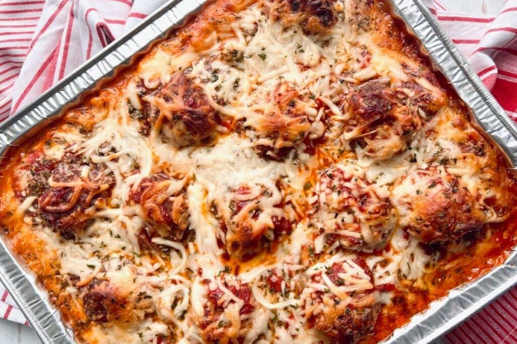 An aluminum baking pan filled with baked ziti with meatballs. The ziti is topped with melted cheese and herbs.