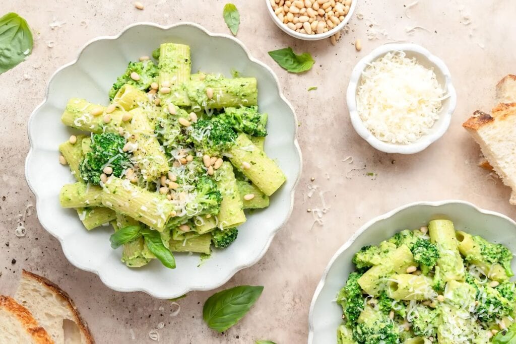 Two bowls of broccoli pesto pasta are shown from above. The pasta is penne, and the sauce is a vibrant green pesto sauce with broccoli florets. The pasta is topped with grated Parmesan cheese and pine nuts. Small bowls of pine nuts and Parmesan cheese are visible in the background. Slices of toasted bread are also visible.