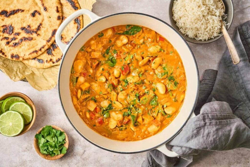 A large pot of butter bean curry is shown from above. Naan bread, rice, lime wedges, and chopped cilantro are visible on the table.
