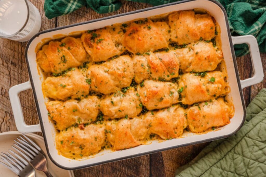 A white baking dish filled with cheesy chicken crescent rolls. The crescent rolls are golden brown and topped with herbs. A glass of milk and forks are visible in the background.