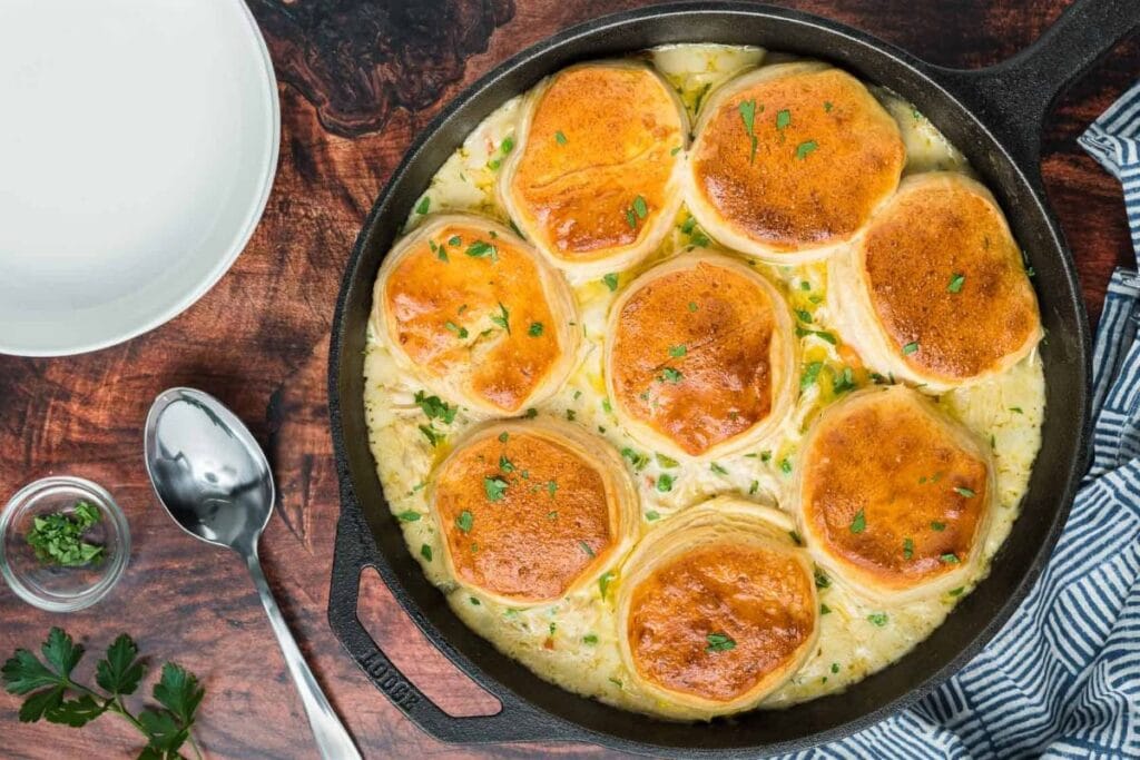 A cast iron skillet of chicken pot pie. The pot pie is topped with golden brown biscuits and sprinkled with fresh parsley. A spoon and small bowl of parsley are visible in the background.