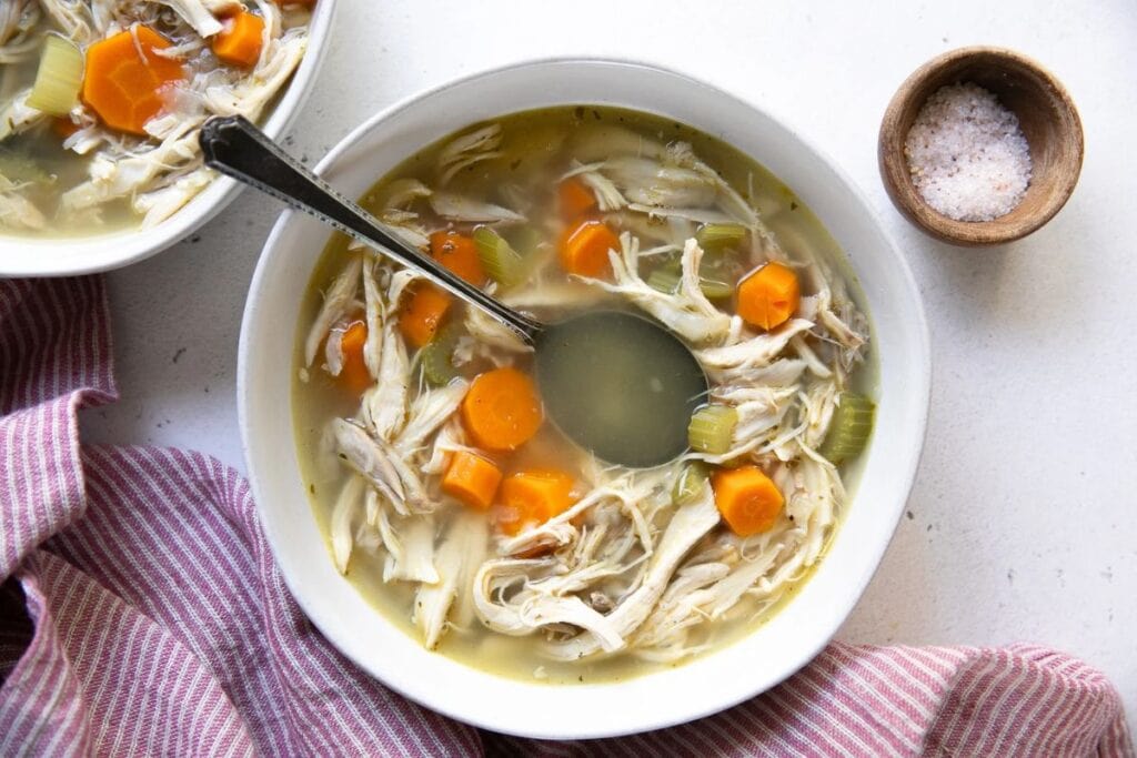 Overhead view of two bowls of chicken soup. The soup is light-colored broth with shredded chicken, carrots, and celery. A spoon is in one of the bowls. A small bowl of salt is in the upper right corner. A pink and white striped napkin is in the bottom left corner.