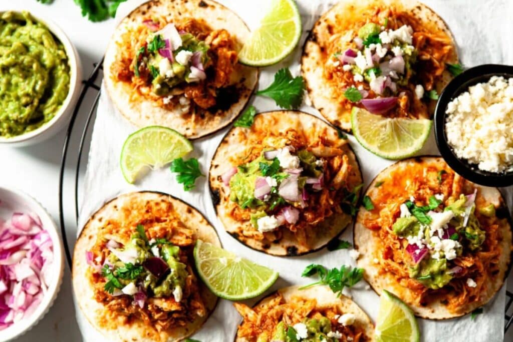 Several small tacos are arranged on parchment paper. Each taco is filled with shredded chicken, guacamole, red onion, and crumbled cheese. Small bowls of guacamole, red onion, and crumbled cheese are also visible. Lime wedges and cilantro sprigs are scattered around the tacos.