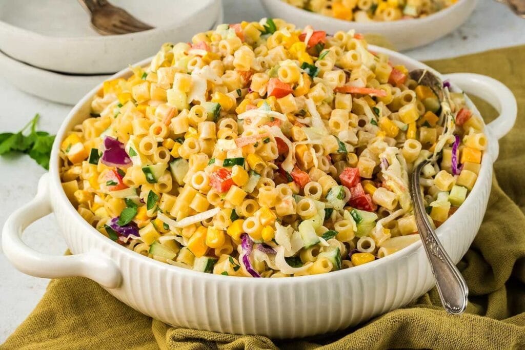 A large white bowl of coleslaw pasta salad. The salad is creamy and contains pasta, coleslaw, corn, and other vegetables. A silver serving spoon is in the bowl.