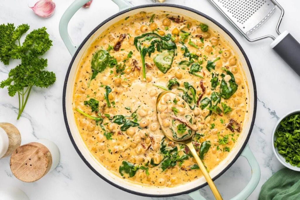 A light green skillet of creamy Tuscan chickpeas with spinach and sun-dried tomatoes is shown from above. A gold spoon is in the skillet. Fresh parsley and salt and pepper shakers are visible on the table.