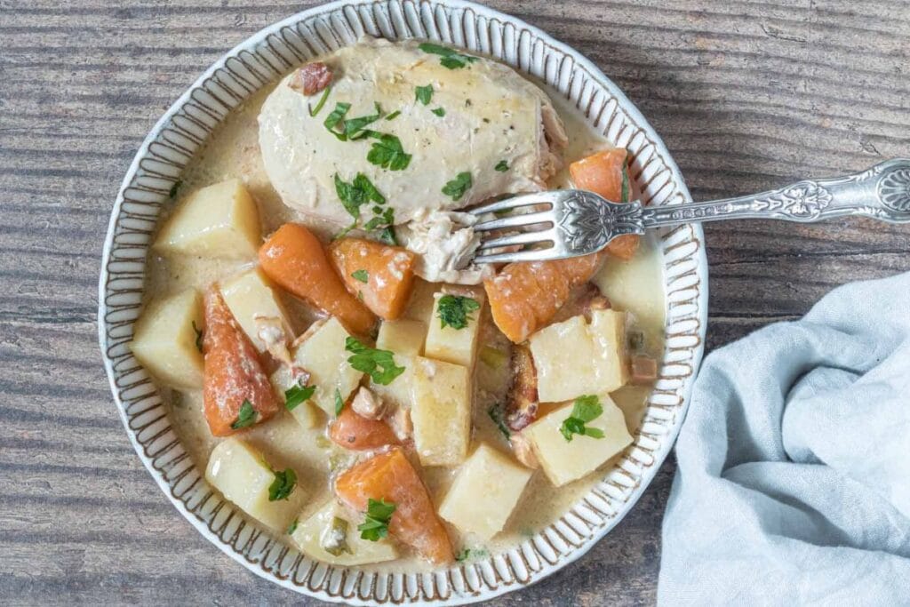 A plate of creamy chicken and vegetables. The chicken is on top of potatoes and carrots in a creamy sauce. A fork is taking a bite of the chicken.