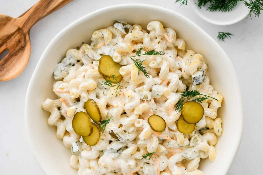 A white bowl filled with pasta salad. The salad is creamy white with green dill and yellow pickle slices.