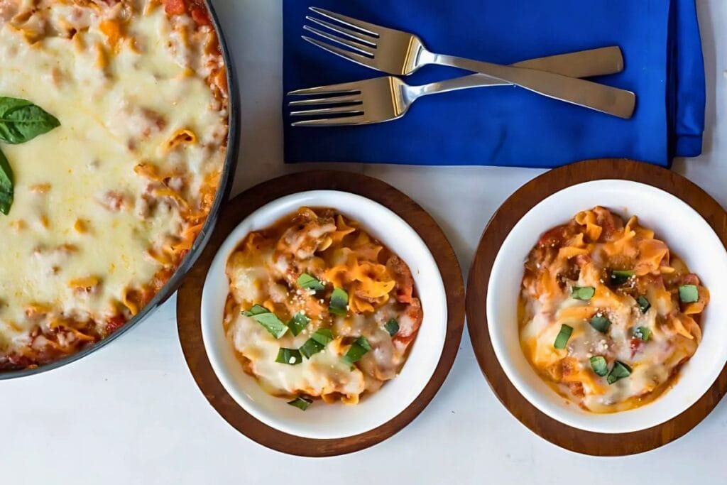 An overhead shot shows a large skillet of baked lasagna and two smaller bowls of the same dish. The lasagna is topped with melted mozzarella cheese and fresh basil. Two forks rest on a blue napkin.