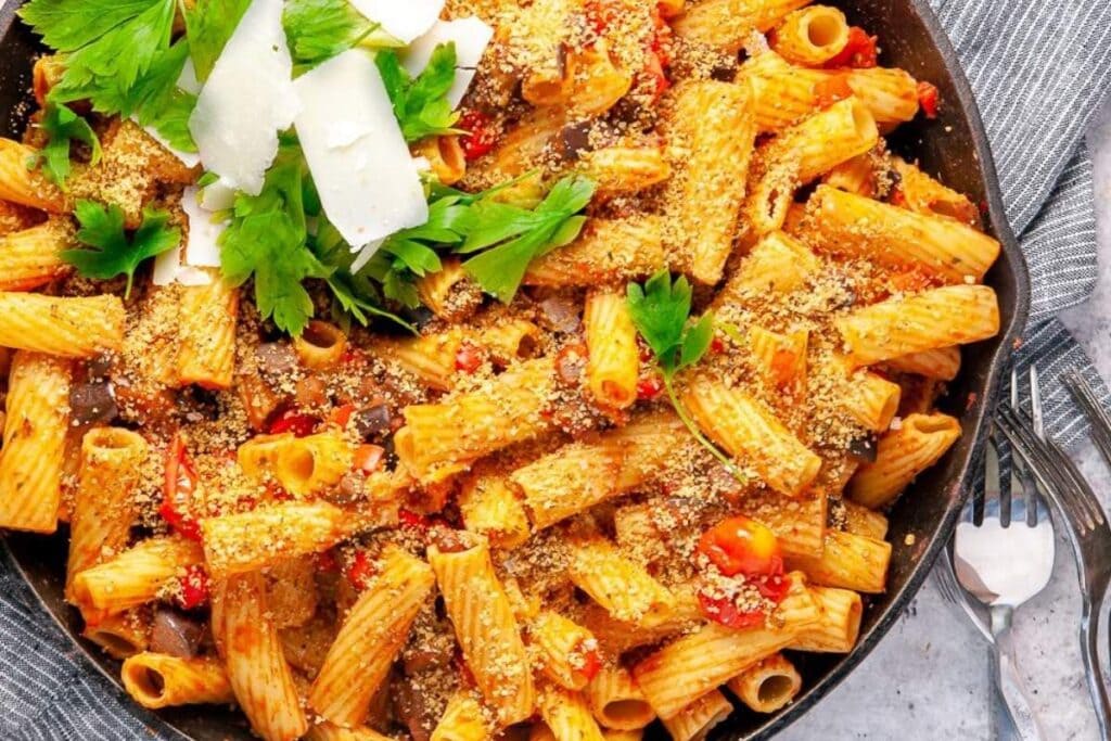 A skillet of pasta is topped with a sprinkle of what looks like breadcrumbs and fresh parsley. Slices of what appears to be parmesan cheese are also on top.