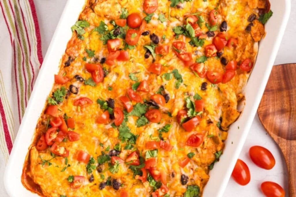 A white baking dish filled with a cheesy casserole. The casserole is topped with chopped tomatoes, cilantro, and black beans. A wooden spoon is next to the dish.