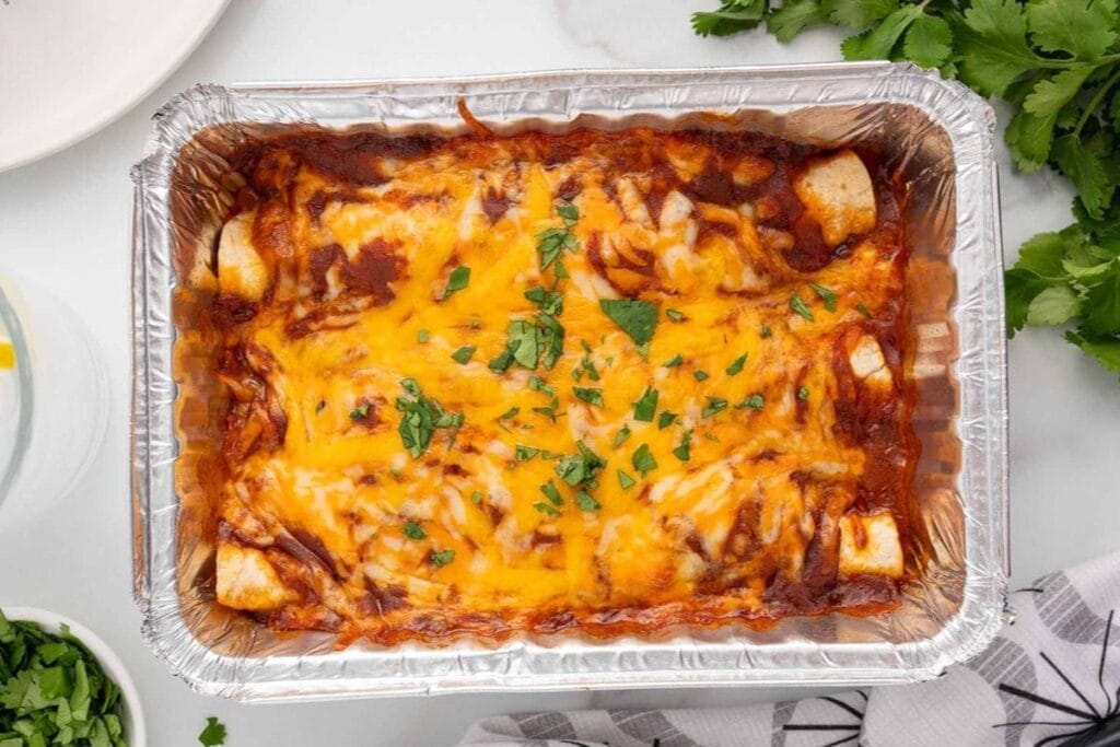 A rectangular aluminum baking pan filled with enchiladas. The enchiladas are topped with melted cheese and chopped cilantro.