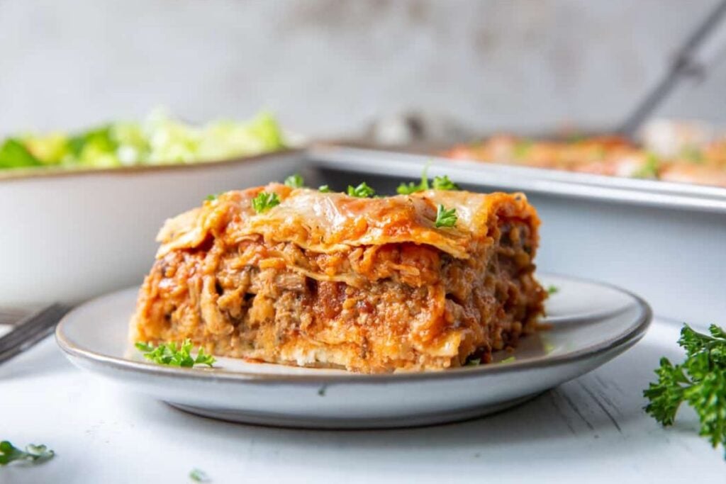 A slice of lasagna on a plate, garnished with parsley. A baking dish of lasagna and a bowl of salad are visible in the background.